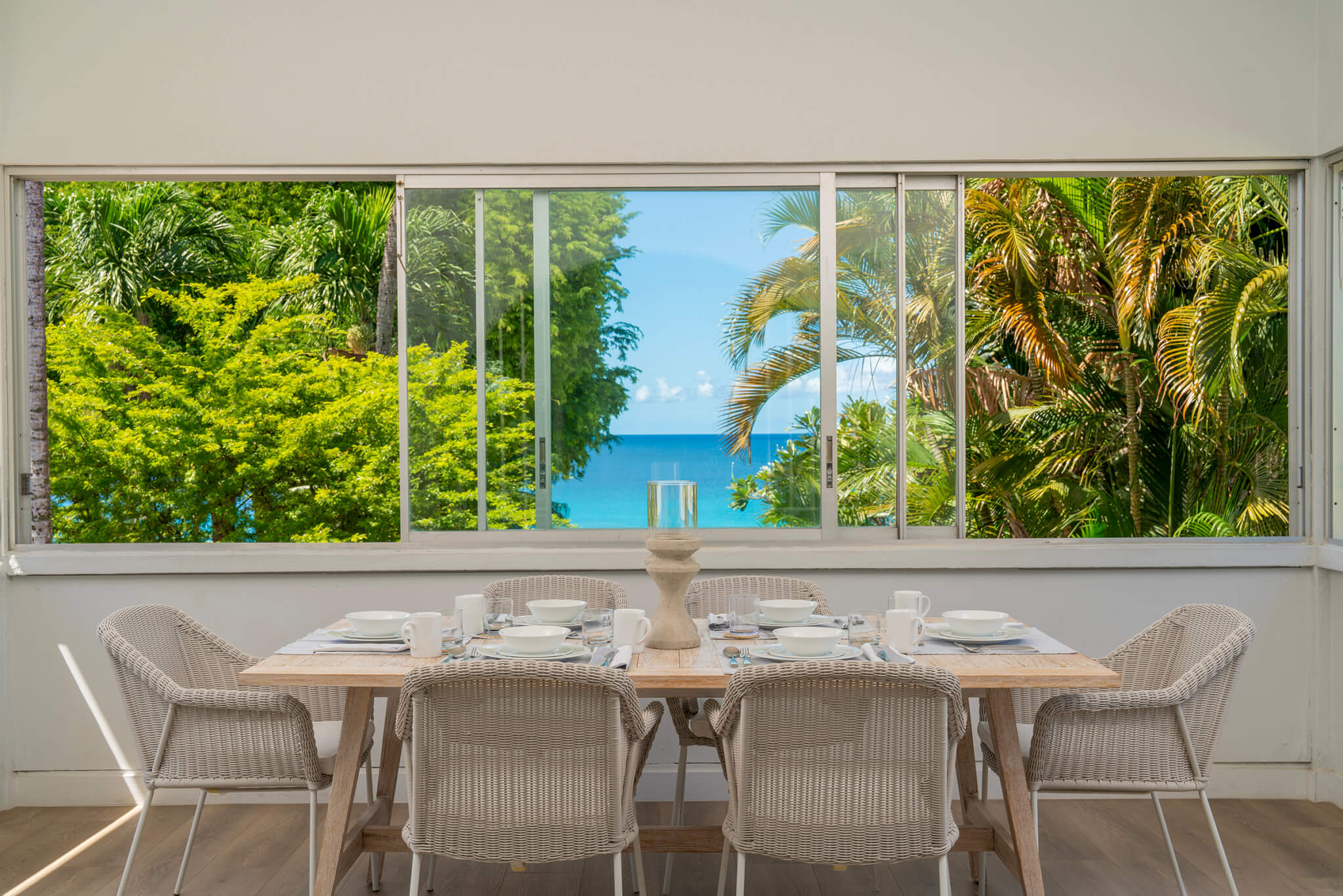 Dining area with a table seating six with a window spanning an entire room looking over turquoise waters and palm trees